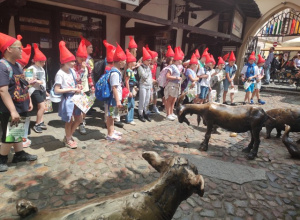 Uczniowie przebrani w czerwone czapeczki krasnali, stoją na Starych Jatkach we Wrocławiu i oglądają stojące obok, naturalnej wielkości figury zwierząt gospodarskich.