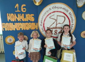 Elegancko ubrani uczniowie pozują przed dekoracyjnym, konkursowym tłem . W rękach trzymają otrzymane dyplomy i nagrody.