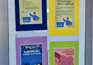 Nowa tablica ogłoszeniowa na szklanych drzwiach naszej szkoły, pełna kolorowych plakatów i ogłoszeń.