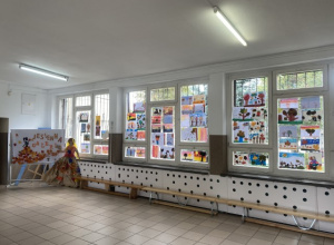 Korytarz szkolny z wyeksponowanymi pracami uczniów przyklejonymi do okien.