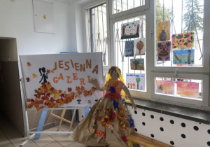 Tablica informująca o jesiennej galerii prac uczniów oraz postać Pani Jesieni wykonana z szarego papieru i kolorowych, papierowych liści.