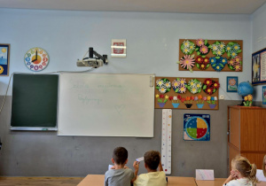 Klasa 1A siedząca w ławkach patrząca się na tablice z napisem “Szkoła Myślenia Pozytywnego”.