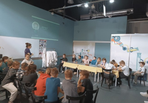 Pani prowadząca zajęcia tłumaczy zagadkę obrazkową uczniom przy stole.