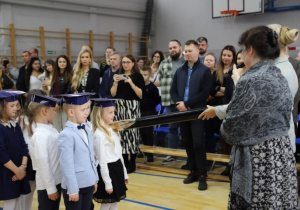 Pierwszoklasiści w strojach galowych w czasie pasowania przez panią dyrektor.