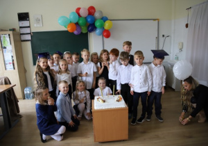 Odświętnie ubrani pierwszoklasiści z wychowawcą stoją wokół wielkiego tortu.