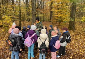 Dzieci wraz z przewodnikiem stoją w grupie. Pani rozdaje im orzechy. Zadaniem dzieci jest schowanie orzechów pod drzewem, a pod koniec wycieczki odnalezienie ich.