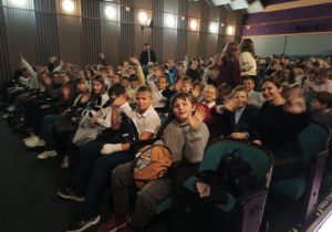 Uczniowie klas 4. siedzą na widowni w teatrze i machają do aparatu.