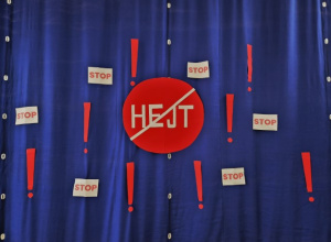 Czerowny znak z przekreślonym napisem hejt zawieszony na niebieskiej kurtynie wraz z napisami “STOP” oraz wykrzyknikami.