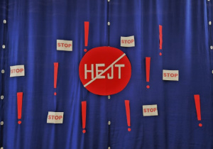 Czerowny znak z przekreślonym napisem hejt zawieszony na niebieskiej kurtynie wraz z napisami “STOP” oraz wykrzyknikami.