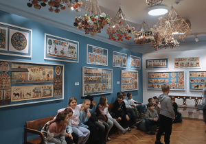 Uczniowie siedzą na ławkach i podziwiają wystawę wycinanek łowickich.