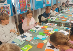 Uczniowie siedzą przy stolikach na których leżą kolorowe kartki papieru do wycięcia wycinanki.