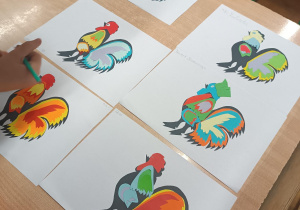 Wystawa na stole gotowych prac – wycinanki łowickiej przedstawiającej kolorowego koguta.