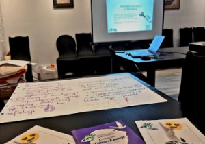 Materiały edukacyjne rozłożone na czarnym stole, w tle widać kolejne slajdy ze szkolenia.