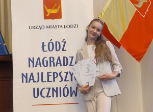 Hania z dumą prezentuje dyplom Stypendium Prezydenta Miasta Łodzi „Mia 100 Zdolnych”, na tle napisu Łódź nagradza najlepszych uczniów.