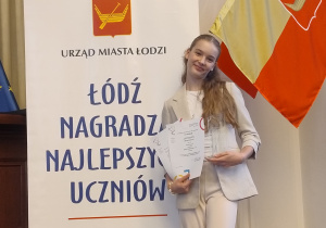 Hania z dumą prezentuje dyplom Stypendium Prezydenta Miasta Łodzi „Mia 100 Zdolnych”, na tle napisu Łódź nagradza najlepszych uczniów.