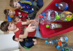 Uczeń klasy 1b przelewa wodę z mniejszej do większej butelki.