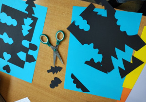 Uczniowie wycinają kształty z czarnego papieru i układają je na niebieskim.