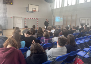 Uczniowie siedzą na krzesłach w sali gimnastycznej i słuchają prelekcji pani mecenas.