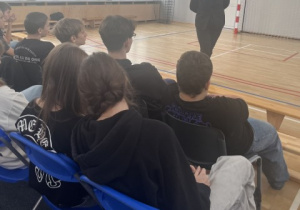 Uczniowie siedzą na krzesłach w sali gimnastycznej i słuchają prelekcji pani mecenas.