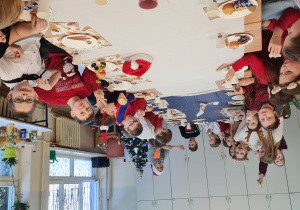 Dzieci siedzą przy stole z białym obrusem i jedzą słodkości oraz owoce.