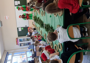 Dzieci siedzą przy stole i wbijają gałązki w wianki.