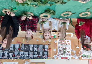 Dzieci siedzą przy stole i wbijają gałązki w wianki.