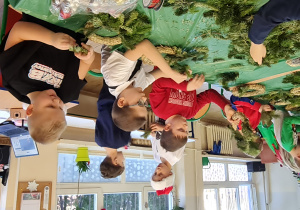 Dzieci siedzą przy stole i wbijają gałązki w wianki.