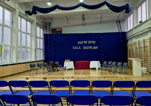 Sala gimnastyczna przygotowana na debatę, z pozostawionymi krzesełkami i gotową dekoracją.