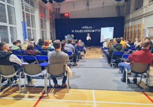 Rodzice siedzą na sali gimnastycznej słuchają przemówienia pani dyrektor.