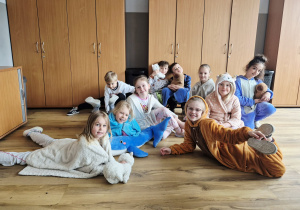 Grupowe zdjęcie dzieci z 1A na końcu sali 116. Dzieci ubrane są w kolorowe piżamki oraz zabrały do szkoły swoje ulubione poduszki, kocyki i zabawki.