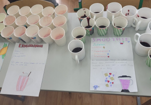 Trzy rodzaje napoi w kubkach ustawione na stole wraz z kartkami zawierającymi niemiecką nazwę, skład i opis wykonania