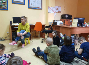 Uczniowie siedzą na podłodze biblioteki i słuchają opowieści pani bibliotekarki