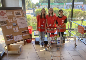 Troje uczniów w czerwonych kamizelkach Caritasu stoją za wózkiem sklepowym wypełnionym produktami sklepowymi