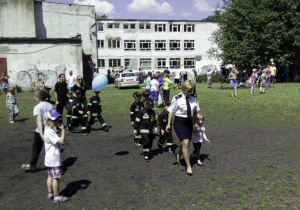 Dzieci ubrane w stroje straży pożarnej maszerują parami przez boisko szkolne