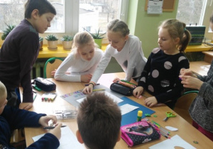 Grupa uczniów przygotowuje plakat na niebieskim brystolu wykorzystując różne materiały i przybory plastyczne.