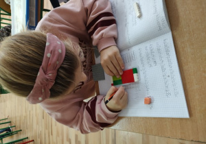 Uczennica zapisuje w zeszycie formułę matematyczną, korzystając z wcześniej ułożonej wieży z klocków.