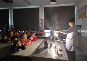 Uczniowie klasy 2 b siedzą w ławkach i oglądają pokaz palenia magnezu przeprowadzany przez ubraną w biały fartuch nauczycielkę chemii.