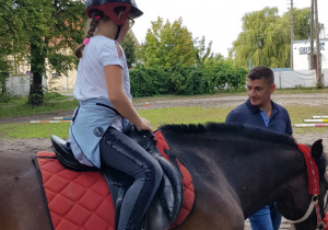 Uczennica ubrana w kask ochronny odbywa przejażdżkę na koniu pod opieką instruktora.