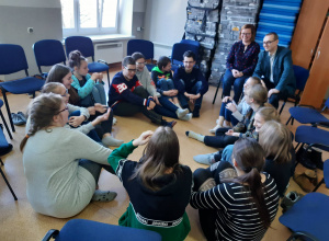 Uczniowie siedzą w kole na podłodze i rozmawiają z parą psychologów.