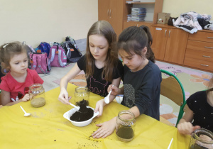 Uczennica napełnia słoik ziemią wsypując ją plastikowa łyżką