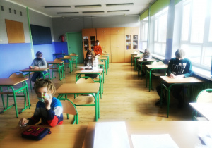 Uczniowie w maseczkach siedzą pojedynczo w ławkach i rozwiązują test.