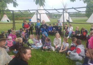 Uczniowie siedzą w kole na tle tipi i słuchają opowieści przewodnika.