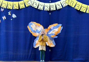 Uczennica podczas recytacji wiersza stoi na tle dekoracji z motylami.