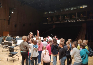 Uczniowie klasy 2B stoją na scenie filharmonii, przyglądając się przychodzącym na próbę muzyką.