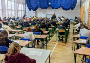 Uczniowie biorący udział w konkursie siedzą w ławkach w sali gimnastycznej.